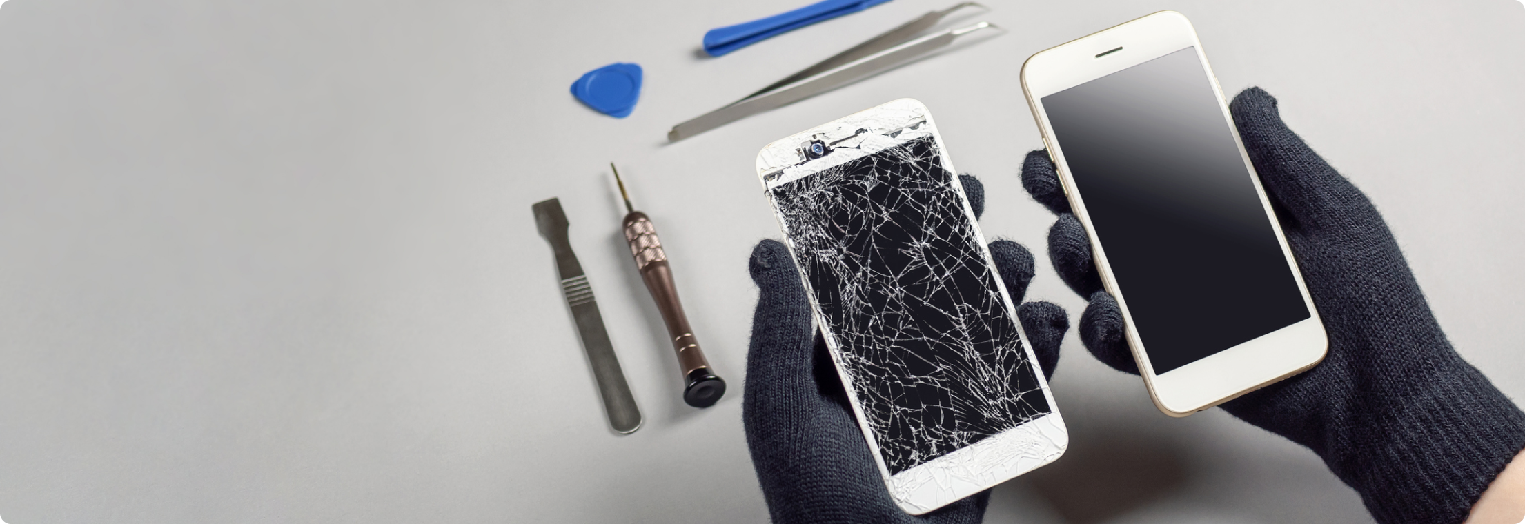 Στη φωτογραφία φαίνονται δυο χέρια με μαύρα γάντια να κρατάνε δυο λευκά κινητά τηλέφωνα, το ένα με σπασμένη οθόνη. Στο φόντο φαίνονται εργαλεία που χρησιμοποιούνται στην επιδιόρθωση κινητων τηλεφώνων.