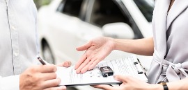Πωλητής παρουσιάζει συμβόλαιο σε αγοραστή με κλειδιά αυτοκινήτου επάνω και το αυτοκίνητο στο φόντο