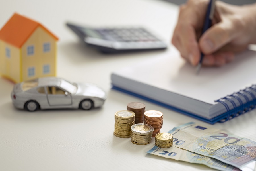 Άνθρωπος σημειώνει σε τετράδιο περιτριγυρισμένο από μια αριθμομηχανή, ένα μοντέλο σπιτιού, ένα μοντέλο ασημένιου αυτοκινήτου και στοίβες από νομίσματα